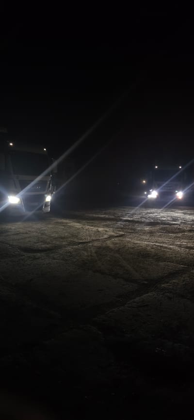 The image shows two trucks parked on a dirt or gravel lot at night. The trucks have their headlights on, illuminating the foggy or dark environment. The truck on the left is closer to the camera and appears to be white. The truck on the right is further away and also appears to be white. The ground is uneven and shows tire tracks. There are light beams from the truck headlights cutting through the darkness and mist. The scene suggests a parking area or a roadside location, possibly a truck stop or a rest area. The specific location in Izmail, Ukraine, cannot be determined from the visual information provided in the image alone.