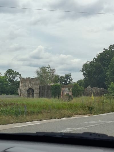 The image depicts a roadside view of what appears to be an abandoned or derelict outdoor exhibit or attraction, possibly in the vicinity of Ravna Gora, Bulgaria. In the foreground, a paved road with a white dividing line is visible, with the edge of a vehicle's dashboard or windshield in the bottom frame. Beyond the road is a field of tall, dry grass interspersed with some green vegetation and wildflowers.
In the middle ground, there are several structures. To the left, a stone-built structure resembling a small fortified gatehouse or tower stands with an arched entrance. It has a crenellated top and appears to be constructed from rough-hewn stone blocks. To the right of this structure, a weathered wooden fence runs horizontally. Beyond the fence, further to the right, are more derelict structures, including what looks like a pinkish building with arched windows and a segment of a broken wall with some decorative elements or sculptures. Scattered amongst these structures are overgrown bushes and trees. In the background, a dense line of dark green trees forms a backdrop against a cloudy, overcast sky. A thin, dark line, possibly a utility wire, stretches horizontally across the upper portion of the image. No people are visible in the scene.