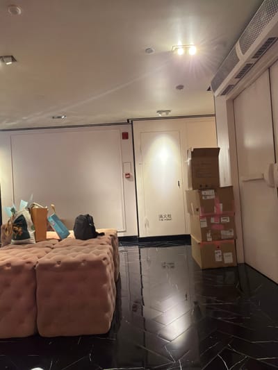 The image depicts an indoor scene with a large, tufted, light pink sofa or ottoman in the foreground. On the sofa, several items are placed, including a dark backpack, shopping bags with various designs, and what appears to be a gift bag. Behind the sofa, there is a large, plain, white wall with two red fire alarm signs. To the right of the wall, there is a white door with Chinese characters "消火栓" and "FIRE HYDRANT" written on it, indicating a fire hydrant. Several stacked cardboard boxes are visible next to this door. The floor is a glossy black tile with a herringbone pattern and white veining, reflecting the overhead lighting. The ceiling is a neutral color with recessed lighting and an air conditioning unit visible on the right side. The scene suggests an indoor waiting area or a room with deliveries.