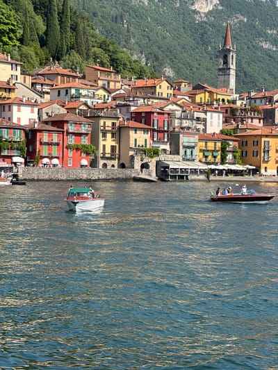 The image depicts a waterfront scene in Varenna, Italy, featuring a town built on a hillside overlooking a body of water. The architecture is characterized by colorful buildings with terracotta roofs, stacked closely together on the steep slope. Prominently visible is a tall bell tower with a red conical spire, a common feature of Italian towns.
In the foreground, the water is choppy, reflecting sunlight and the surrounding scenery. Two boats are present on the water. The boat closer to the viewer on the left appears to be a white motorboat with a green canopy and several individuals on board. Another boat, a classic wooden motorboat, is positioned to the right, also carrying passengers.
Along the waterfront, there are buildings with awnings and outdoor seating, suggesting a commercial area with restaurants or cafes. People are visible walking along the promenade and seated at tables. Trees and lush greenery cover the hillside behind the town, adding to the picturesque setting. The overall impression is a vibrant and scenic Italian lakeside town.