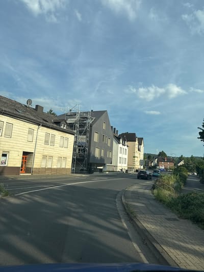This media file captures a street view in Wetzlar, Germany. The scene is dominated by a street lined with various buildings, ranging from older, traditional structures to a more modern, gray building with a dark facade. Scaffolding is visible on the modern building, suggesting ongoing construction or renovation work. The street itself is asphalt with clear white lane markings.

On the sidewalk to the right of the street, there is a paved walkway. Trees and vegetation are present along the sidewalk and in the background, indicating a residential or urban area with greenery. Several cars are parked or driving on the street, including a black SUV and a blue hatchback. The sky is visible with scattered clouds against a bright blue background, suggesting it is daytime, likely mid-morning or afternoon, with fair weather.

One of the buildings has a visible house number "19" and an orange-framed window. A sign is attached to the scaffolding, but the text on it is not clearly legible. The overall impression is a typical European street scene on a pleasant day, with a focus on architecture and urban life. No people are prominently featured in the image. The perspective suggests the image was taken from a moving vehicle, looking out at the street.