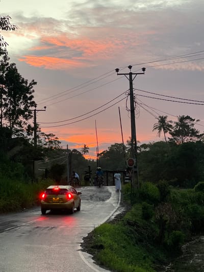 The image shows a road scene in Kamburugamuwa, Sri Lanka. A car with its brake lights illuminated is driving on a wet, winding road. Two cyclists are visible ahead of the car. A railway crossing gate is lowered across the road. Overhead power lines extend across the scene. Palm trees and other vegetation are visible. The sky is illuminated by a sunset with orange and grey clouds.