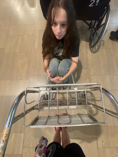 An overhead, wide-angle shot captures a scene at an airport in Israel. A young female, with long brown hair and bangs, is positioned centrally, squatting or sitting on a low stool. She wears a black graphic t-shirt, light blue jeans, and dark sneakers, with her arms wrapped around her knees. A colorful bracelet is on her left wrist, and a thin bracelet on her right. She gazes upwards towards the camera with her mouth slightly open. Directly in the foreground, partially obscuring the lower half of the girl, is a chrome-plated metal luggage cart with a flat bottom and basket-like upper shelf. The pink-shoed feet and lower legs of the person holding the camera are visible behind the cart, along with a portion of a patterned garment and a dark bag. In the background to the right of the girl, a black wheelchair with the white number "21" on its backrest is partially visible. The floor is covered with light-colored square tiles.