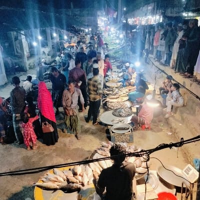 The image displays an active night market scene in Lalmohan, Bangladesh. A dense crowd of individuals, predominantly adult males, occupies an unpaved outdoor market area. Numerous vendors are seated on the ground or low stools behind large circular metal platters and mats, which display various species of fish.

In the foreground, a person is positioned with their back to the camera, attending to a substantial quantity of fish arranged on a yellow circular mat. Adjacent objects include a blue plastic crate and a weighing scale. Towards the mid-left, a woman wearing a red hijab and dark attire stands alongside a young child dressed in a pink garment. A man in a light-colored shirt and striped lungi is centrally located in the mid-ground.

On the right side, several individuals, including children, are seated on a raised concrete platform, observing the market activity. Black electrical utility wires traverse the upper foreground. The scene is intensely illuminated by multiple artificial light sources, including bare bulbs, creating a high-contrast visual environment. Buildings and rudimentary market stalls are discernible in the background. The depicted activity indicates ongoing commercial transactions within a bustling public space.