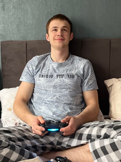 An image, rotated ninety degrees counter-clockwise, depicts a young man relaxing indoors while playing a video game. The man, with short brown hair and a light complexion, is positioned from the waist up, facing towards the right side of the frame with a slight smile on his face. He is wearing a light-colored, possibly light blue or grey, t-shirt with a repeating pattern of thin, wavy lines. On the front of his t-shirt, the text "UNIQUE FOR YOU" is clearly visible in a darker font, arranged vertically.
He is holding a black video game controller, which appears to be a PlayStation controller, with both hands. The light bar on the controller is illuminated with a bright blue glow, indicating it is powered on and in use. His right leg is visible, covered by black and white plaid pajama bottoms, suggesting a casual, at-home setting. Another part of a similar controller or device is partially visible on the far right edge of the frame, hinting at potential multi-player activity or a second controller.
The man is reclining on what appears to be a bed or a comfortable couch, with a dark brown headboard or backrest behind him. A white pillow is visible beneath his left arm and torso. The wall behind him has a dark, possibly charcoal grey or dark blue, textured finish. The overall impression is one of leisure and enjoyment, likely at home.