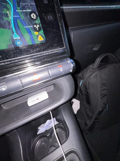A center console of a car shows a navigation screen displaying a map, next to which is a vertical control panel with buttons. A small compartment holds a white USB drive and a cable leading to cup holders containing folded paper. A black bag rests above the console, partly obscuring a section of the dashboard. The surfaces appear slightly dusty, suggesting recent use.