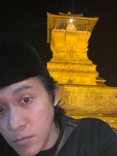 A close-up, low-light selfie captures an individual against the illuminated clock tower of the Great Mosque of Kauman in Yogyakarta, Indonesia. The individual, appearing male, occupies the lower left half of the frame, wearing a black peci and a dark garment, with a dark bag strap visible on their right shoulder. Their face is illuminated by a direct light source, showing distinct features and some facial redness. Their dark hair is visible below the peci. The background features a prominently illuminated, multi-tiered yellow-orange stone or brick clock tower, a significant architectural element of the mosque. A white-faced clock with black hands is visible on the upper tier of the tower, indicating the time as approximately 11:25. The sky above and around the tower is dark, suggesting nighttime.