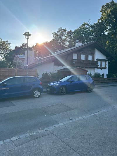 This image depicts a street scene in Höhenkirchen-Siegertsbrunn, Germany. The primary subjects are two blue cars parked on an asphalt road. One car, a smaller hatchback, is in the foreground, while a larger car, possibly a sedan, is parked behind it. Both cars are facing away from the viewer, with their license plates visible on the rear of the larger car.

The setting includes a traditional Bavarian-style house with white walls, dark trim, and wooden balconies. Mature trees with lush green foliage surround the house, and a wooden fence runs along the property line. The sun is low in the sky, casting long shadows and creating lens flare across the image, suggesting it is either early morning or late afternoon. The sky is clear and blue. A street lamp with a distinctive dual-headed design is also visible. The overall impression is one of a peaceful residential area bathed in the warm light of the sun. There are no people visible in the image. The visible text on the license plate of the larger car is "M-HK 395".