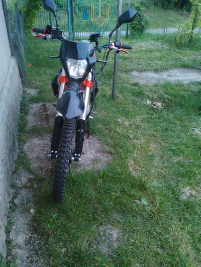 The image shows a black and red dirt bike parked outdoors on a patch of grass next to a concrete wall and a chain-link fence. The motorcycle is the primary subject, positioned centrally in the frame and viewed from a slightly low angle. Its front wheel, handlebars, and headlight assembly are clearly visible. The bike appears to be new or well-maintained. The background includes green grass, some dirt paths, and vegetation, with a glimpse of a blue fence in the distance. The overall setting suggests a residential or backyard area. No people are present in the image. The location is identified as Berezynka, Ukraine.