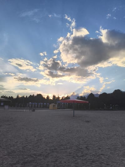 The image shows a sandy beach area with a line of trees in the background. The sky is partly cloudy with the sun setting behind the clouds, casting rays of light. In the foreground, there is a red umbrella and a blue pole. Further back, there are several small structures, possibly kiosks or changing rooms, and a fence. The area appears to be a public space, possibly a recreational area or park in Bayet, Kyrgyzstan. There are no people clearly visible in the image. The overall scene depicts a serene, late afternoon or early evening atmosphere.