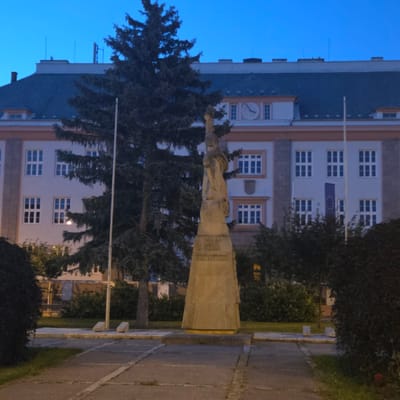 The image shows a monument in the city of Piešťany, Slovakia, located in front of a building. The monument is a stone statue of a figure in a robe, standing on a pedestal. The pedestal has inscriptions on it, though they are difficult to read. Behind the monument is a large building with many windows and a dark roof. A large evergreen tree is situated to the left of the monument, partially obscuring the building. The sky is a deep blue, suggesting it is dusk or dawn. The ground in front of the monument is paved with concrete slabs.