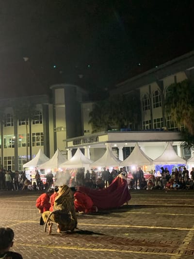 A night performance takes place in front of a large building in Kecamatan Batu, Indonesia, located at coordinates -7.867, 112.514. Five white tent canopies are set up in front of the building. A person wearing a red and gold lion costume performs a dance in the street, their tail dragging behind them. Other performers are wearing red clothing. Spectators are seated on the ground, watching the performance. There are people in the background standing and sitting, some holding drinks. The building is lit with bright lights, and the street is paved with brick. There is smoke coming from the ground near the performers.