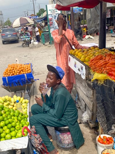 This image captures a street market scene in Langbasa, Nigeria, under an overcast sky. In the foreground, a barefoot male, dressed in a dark green traditional Nigerian top and trousers and a backward blue baseball cap, sits on a black drum labeled "Valu Paints." He is drinking from a clear plastic water sachet with his eyes partially closed, while holding a dark smartphone in his left hand.

To his immediate left, a white display table holds a large quantity of green, round fruits, and striped yellow-green fruits, with a plastic water bottle visible among them. A folded, colorful umbrella with a red handle rests near this display. To the left and slightly behind him, a blue plastic crate filled with numerous small orange fruits rests on a metal wheelbarrow.

Behind the sitting individual, a standing male, wearing a patterned light orange traditional Nigerian garment and a dark shoulder bag, stands behind a wooden fruit stall. He has his right hand raised to his face, partially obscuring it. The stall, covered by a red and white striped canopy, displays a large pile of yellow-orange fruits (likely oranges) and a prominent pile of orange carrots. Below this stall, small red tomatoes are visible in metal buckets.

In the mid-ground, a dark blue SUV is parked on the left side of the unpaved street. Further back, another individual is seen pushing a small cart laden with items. Various other market stalls, umbrellas, and people are visible along the street. A green and white sign for "Furri Classic Schools" is mounted on a pole, and overhead power lines traverse the scene. A water tank on a stand is also visible in the background.