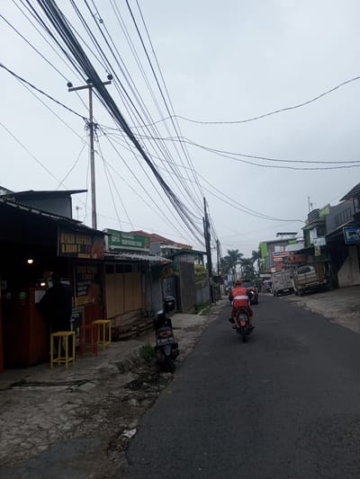The scene depicts an urban street in the city of Houaythat, Laos, under an overcast sky. A paved, dark grey asphalt road occupies the center, curving gently to the right in the distance. On the left, a single-story building with a dark corrugated metal roof houses two businesses: "AYAM GEPREK H.ODING S" (Smashed Chicken H. Oding S) and "KAYLA SEDIA ANEKA BUAH-BUAHAN SEGAR" (Kayla Provides Various Fresh Fruits), indicated by visible signage. One person in a dark shirt is observed behind a counter inside the "Ayam Geprek" stall. Two yellow stools and one red stool are positioned on the cracked concrete sidewalk in front of the stall. A black scooter, with license plate "F 6307 YY", is parked on the sidewalk. A tall utility pole stands on the left, supporting a dense, complex array of black electrical and communication wires spanning horizontally and vertically across the scene. On the road, a person wearing a red jacket with "TRANSPORTER" text is riding a red motorcycle away from the viewer; its license plate "2893 V" is partially visible. Further down the street on the right, multiple buildings, some painted green and grey, line the road, displaying various commercial signs, including "Alfamart" and "J&T EXPRESS" on a white truck. Palm trees are visible in the background between buildings. Other vehicles are present further along the street.