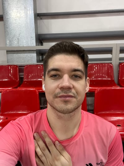 A selfie captures a man in a pink athletic shirt, seated in a red stadium seat. He has short, dark hair, light skin, and a trimmed beard. An earpiece is visible in his ear. His hand rests on his chest. Rows of empty red seats fill the background, alongside gray metal railings and a white wall, suggesting an indoor stadium setting. The image appears to be slightly rotated.