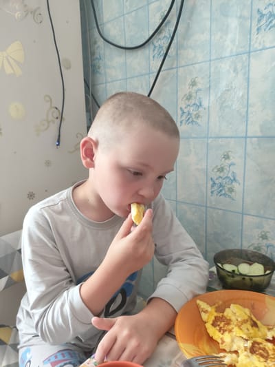 A young boy with short brown hair is sitting at a table and eating a piece of food. He is wearing a long-sleeved gray shirt and pajama bottoms with a pattern of vehicles. In front of him is an orange plate with omelets and a fork. To his right is a bowl of sliced cucumbers. The background consists of a wall with a tiled pattern and decorative wallpaper. The scene appears to be indoors, likely a kitchen or dining area. There is no discernible information in the image to indicate the location is Askiz, Russia.