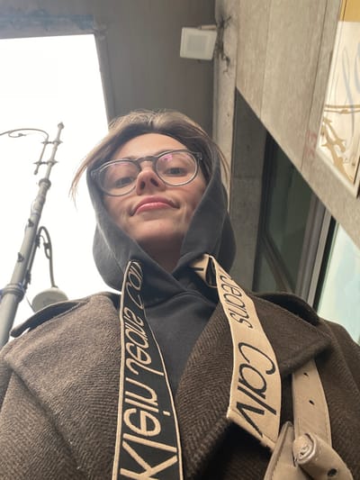 A low-angle portrait captures an individual, likely female, positioned outdoors in Scafati, Italy. The subject wears clear-framed eyeglasses, a dark grey hooded sweatshirt with the hood raised, and a brown tweed-patterned overcoat. A beige strap displaying "Calvin Klein Jeans" in black text diagonally crosses the upper torso. The individual features dark hair framing the face and a slight smile, with direct eye contact towards the camera. The overhead environment consists of an overcast sky. To the left, a dark, multi-tiered street lamp pole is partially visible. To the right, a light-colored building facade features a small white square fixture mounted high, and a reflective surface below, suggesting a window or storefront with indistinct reflective elements.