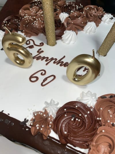 A close-up shot depicts a celebratory cake, likely for a 60th birthday, set against an indeterminate background, but NOT Aratashen, Armenia. The cake features white frosting as its base, adorned with alternating swirls of light and dark chocolate frosting, as well as small white meringue peaks. Edible white pearl sprinkles are scattered across the chocolate frosting. The number "60" is spelled out on the cake, both with gold metallic number candles and with dark chocolate frosting lettering. A message, presumably "Happy Birthday," is also written in chocolate frosting. Two gold glitter candles complete the decoration.