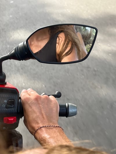 An overhead, eye-level shot shows a person's hand on the handlebar of a red scooter. The hand, with visible hair, is wearing a thin bracelet. The scooter's controls include a red button and a black switch. A side mirror reflects the shoulder and hair of the rider, who is wearing a black top. The background is a gray asphalt road. The location is Midigama, Sri Lanka.