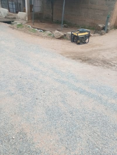 A yellow and black portable generator is situated on a dirt ground next to a brick building in Zinna, Nigeria. The generator is framed by a metal roll cage and has a black handle on top. Adjacent to the generator are large rocks and a metal pole. In the background, the building has a corrugated metal roof supported by a metal frame. A gravel road extends into the foreground, with patches of grass and debris visible along the edge of the road near the building. The scene appears to be an outdoor area, possibly a yard or alleyway, in a residential or commercial district.