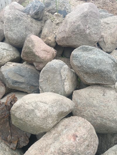 The image shows a pile of various sized, mostly grey rocks. One rock appears to have a pinkish hue. Partially visible on the lower left is some rusted metal. The location is unspecified, though the request mentions Яльгелево, Russia. The image contains no people, actions, or interactions. The environment is natural.