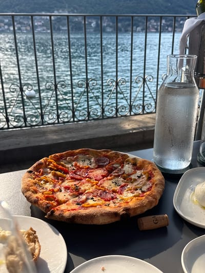An overhead shot shows a pizza on a table in front of a railing overlooking Lake Como in Pognana Lario, Italy. The pizza is on a white plate and has pepperoni and peppers on it. A bottle of water is to the right of the pizza. In the background, the lake is visible with a white buoy in the water.