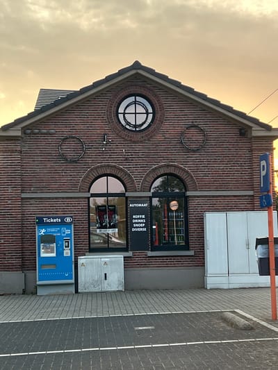 The image shows the exterior of a brick building, likely a small commercial establishment or service point, in Zichem, Belgium. The building has a distinctive gable roof and is adorned with circular decorative elements. Two arched windows are visible on the brick facade, one round and one semicircular.

In front of the building, there is a blue ticket vending machine with the text "Tickets" and a "B" logo, suggesting it is for public transport or a similar service. Next to the ticket machine is a grey electrical cabinet. Above the windows, a sign with a blue "P" symbol indicates a parking area.

Through one of the windows, a display with the text "AUTOMAT", "KOFFIE", "DRINKS", "SNOEP", and "DIVERSE" is visible, indicating that the establishment offers coffee, drinks, snacks, and other items. A clock is also visible inside the window display.

The time of day appears to be late afternoon or early evening, with warm, soft light illuminating the sky, which is partly cloudy. The overall atmosphere is calm and functional, with no people actively engaged in any discernible activity in the frame. The setting seems to be an outdoor area next to a paved walkway or road.