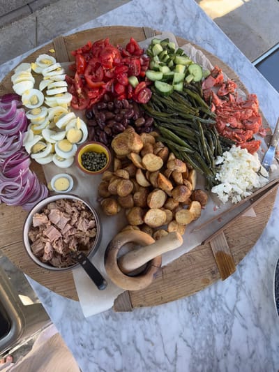 This image displays a large, circular wooden serving board placed on a white marble-patterned table in an outdoor setting, likely a patio, in Area B (Shawnigan Lake), Canada. The board is covered with crumpled parchment paper and features a variety of food items arranged for serving.

From the bottom left, arranged clockwise, the board holds: thinly sliced red onions; vertically stacked halves of hard-boiled eggs; a small, yellow-rimmed ceramic bowl containing green capers; a pile of roughly chopped red tomatoes; a mound of dark, pitted olives; a line of cooked green beans; a pile of diced green cucumbers; a heap of flaked, reddish-orange cooked salmon; a mound of crumbled white feta cheese; and a large central pile of small, golden-brown roasted potatoes. A dark-colored ceramic bowl containing flaked canned tuna is positioned on the lower-left of the board.

A wooden mortar and pestle set is placed on the parchment paper on the lower-right side of the board. Serving utensils visible include a metal fork with a wooden handle near the salmon, a pair of wooden tongs near the feta cheese, and a small black-handled knife or spreader next to the tuna bowl. No individuals are present in the image, and no active interactions are depicted.