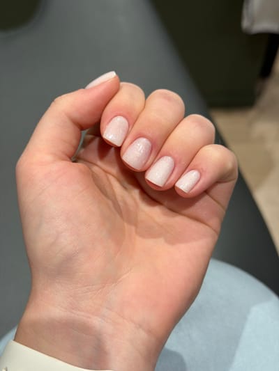 The image shows a close-up of a person's left hand, displaying manicured fingernails painted a light, shimmery pink. The hand is resting on a gray surface, possibly a piece of furniture, with a blurred background that appears to be an indoor environment. The lighting suggests an artificial light source. There are no discernible landmarks or specific architectural features visible that would allow for the identification of a location within Fredrikstad, Norway.