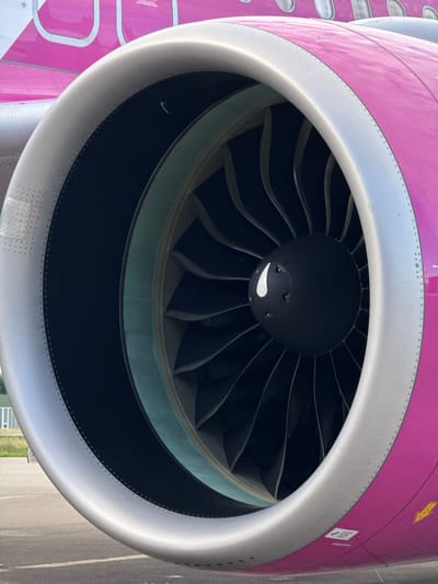 The image shows a close-up view of a jet engine on the exterior of an aircraft. The engine nacelle is predominantly colored in a vibrant pink, with a silver-colored lip around the intake. Inside the intake, visible fan blades are present, with a black spinner at the center featuring a white graphic element. The aircraft is parked on an asphalt surface, likely an airport tarmac, with a hint of grassy ground and a building in the background, suggesting an outdoor setting. There are no people or actions depicted. The location is specified as Künersberg, Germany.