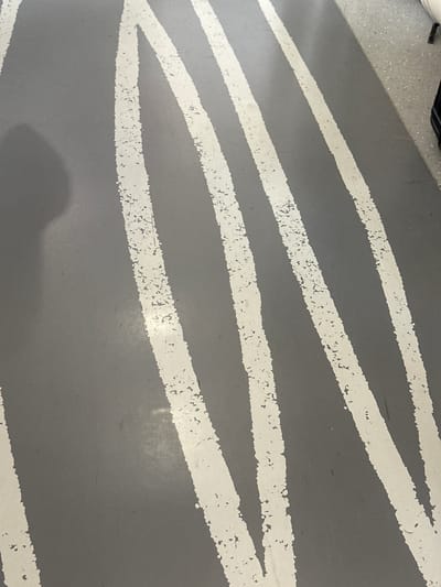A close-up, high-angle shot of a gray floor with several thick, white, diagonal stripes. The stripes are uneven, with a speckled texture, and appear to be painted or adhered to the surface. The floor has a matte finish. A faint shadow of a person is visible on the left side of the image. The context suggests an indoor environment, possibly a public space like a gymnasium, parking garage, or commercial area. Without further information, pinpointing the exact location within Guangming District, China, is not possible, but the scene depicts a common urban flooring pattern.