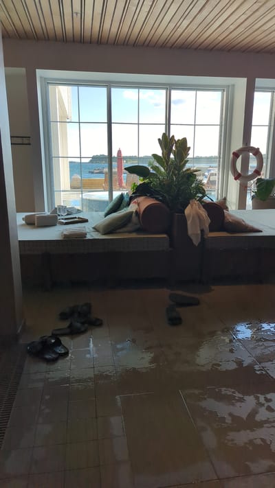 The image depicts an indoor scene, likely a spa or relaxation area, with a large window overlooking a body of water and a distant shoreline. The floor is tiled and wet, with several pairs of dark-colored flip-flops scattered across it.
In the foreground, on the wet tiled floor, are multiple pairs of dark-colored flip-flops, some appearing to be submerged in puddles of water.
The midground features a raised, cushioned seating area with several pillows and folded towels. A large potted plant with green foliage is situated on the seating area.
The background is dominated by a large window with multiple panes, offering a view of a bright, outdoor scene. Outside, a body of water is visible, with a distant shoreline featuring buildings and vegetation. The sky is blue with scattered clouds. A red life preserver ring with white accents is mounted on a white pole to the right of the window.
The environment appears to be a well-lit indoor space with a wooden plank ceiling. The wet floor suggests recent use of the area, possibly for swimming or showering. The overall impression is one of a relaxed, possibly resort-like setting. The scene is consistent with a location in Larkollen, Norway, known for its coastal environment and recreational facilities.
