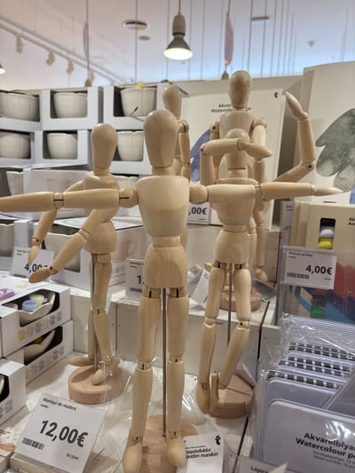 The image is a close-up shot of a retail display featuring several wooden artist mannequins. The mannequins are light-colored wood and are posed in various dynamic positions, with articulated limbs. They are positioned in the foreground and middle ground of the image.
In the background, shelves are stocked with white ceramic bowls and other products, suggesting this is likely a store that sells art supplies, home goods, or a combination of both. A sign partially visible in the background reads "Akvan Watercolor".
There are several price tags visible. One tag reads "Maniquí de madera Hobby 12,00€". Another tag reads "Pintura acrílica Hobby 4,00€". There is also a tag that says "Taza flor, D Hobby 4,00€".
The lighting appears to be from overhead fluorescent fixtures. The overall environment is a retail space.
While the image is taken in Orihuela, Spain, the exact location within the city is not discernable from the visual information provided. There are no people visible in the image. The focus is on the inanimate objects for sale.