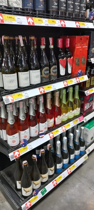 This image captures a well-stocked aisle within a supermarket or liquor store, likely located in Moo 1, Thailand. The scene displays multiple shelves filled with various bottles of wine and sparkling wine, neatly arranged in vertical and horizontal rows.

The products include a range of wines such as red wines (e.g., "CABERNET S" from "RICASON" and "ROBERT MO"), white wines ("ROTHBURY ESTATE"), rosé, and sparkling wines, some of which appear to be fruit-flavored like "fresh PEACH." Prominently featured brands include "RICASON," "ROBERT MONDAVI," "ROTHBURY ESTATE," and "JACOBS CREEK."

White price tags with red directional arrows are affixed to the shelves, indicating prices and product locations. These tags display prices such as 1,945.00, 845.00, 705, 687, 804, 379.00, 798, 540, 214, 528, 378, and 1,485, which, given the location, are likely in Thai Baht. Some price tags also feature QR codes. Small circular flags are visible on some bottles, with an Australian flag seen on a Jacob's Creek bottle and a blue, white, and red vertical-striped flag, possibly French, on a sparkling rosé bottle. The overall setting is an indoor retail environment, designed for product display and sale. No people or specific activities other than product merchandising are depicted.