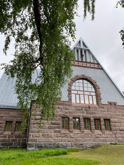 The image shows the exterior of a building, likely a historical structure, made of rough-hewn reddish-brown stone blocks. The building has a steep, grey, metal-clad roof that tapers to a point. A large, arched window with a wooden frame and many panes is centrally located on the upper part of the stone wall. Below this, several smaller rectangular windows with diamond-shaped patterned glass and wooden frames are visible. The building is partially obscured by the branches and green leaves of a tree in the foreground, which hangs over the scene from the left. The sky is overcast and grey. The ground in front of the building is covered in green grass. Based on visual cues and architectural style, this building is identified as the Melnikovo Power Plant Museum, located in Melnikovo, Russia. There are no people or animals visible in the image.