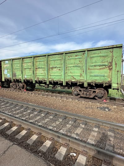 The image depicts a green freight car on a railway track, likely located in or near Кечёво, Russia. The rail car shows signs of wear and age, with visible rust and markings. The railway track is laid with gravel and concrete sleepers. The sky is partly cloudy.