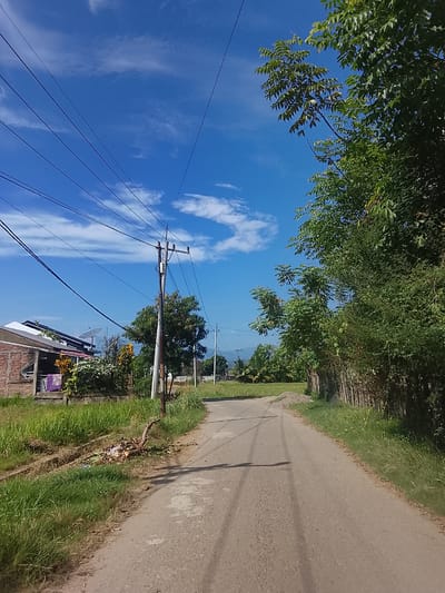 The image depicts a paved road in a rural area, likely Geuleudah, Indonesia, under a bright blue sky with scattered white clouds. On the left side of the road, a low-rise building with a brick facade is visible, with laundry hanging on a line. Adjacent to the building is a patch of tall green grass and a drainage ditch containing some debris. Overhead, numerous power lines stretch across the sky, supported by a utility pole. To the right of the road, a dense line of trees and vegetation forms a natural barrier, with a wooden fence partially visible. The road itself shows tire tracks and is flanked by more green grass and sparse vegetation. In the distance, more trees and what appears to be a rice field can be observed, suggesting a predominantly agricultural or semi-rural landscape. The scene is devoid of any people or visible activity.