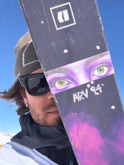 A person wearing sunglasses and a knit hat is holding a snowboard in front of their face. The snowboard has a purple and black design with a pair of eyes and the text "ARV 94". The person is wearing a black hoodie with a white jacket underneath. The background is a clear blue sky. The location is likely Saint-Martin-de-Belleville, France, considering the snowy environment suggests a ski resort area.