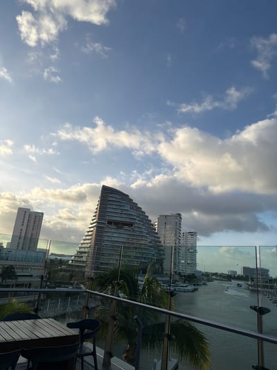 The image shows a view from a balcony overlooking a marina in Cancun, Mexico. In the foreground, there is a wooden table and four chairs on a balcony with glass railings. Beyond the balcony, a canal or river is visible, lined with boats and modern buildings. The architecture of the buildings is contemporary, with many featuring glass facades. The sky is partly cloudy with dramatic lighting, suggesting it might be late afternoon or early morning. The overall scene depicts a vibrant, urban waterfront environment in Cancun.