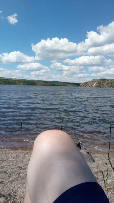 The image shows a person's leg in the foreground, with a body of water and a landscape in the background. The person appears to be sitting by the water, with fishing rods set up. The water is rippling and the sky is blue with white clouds. The landscape in the distance consists of rolling hills covered in vegetation and some rocky outcrops. The ground in the foreground is covered with small stones and pebbles. The overall impression is of a peaceful outdoor scene, likely a fishing spot.