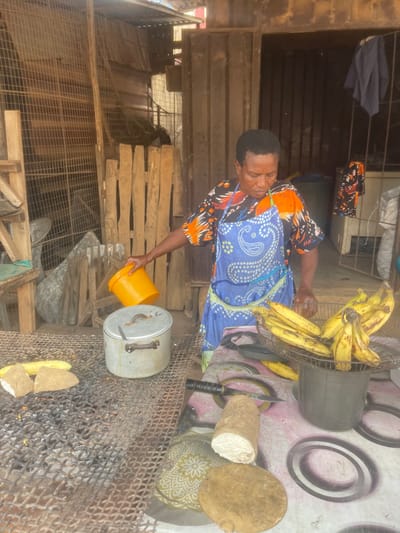 The image captures a scene featuring a woman engaged in food preparation, likely outdoors in a market or roadside stall setting. The image is oriented vertically, so describing it as if it were rotated 90 degrees clockwise would present a more natural viewing perspective.
In the center, a woman with dark skin and short, dark hair is seen from the chest up, looking downwards towards an aluminum pot. She is wearing a patterned short-sleeved top with orange, black, and white designs, and a blue apron with a white paisley-like pattern tied around her waist. Her right arm is extended, holding a bright yellow plastic bucket, from which she appears to be pouring liquid or contents into the open aluminum pot below. The pot has a lid with a handle resting beside it on the mesh surface.
To the right of the woman and slightly behind her, a large bundle of ripe yellow bananas is piled within what looks like the cage or guard of a standing fan, perhaps repurposed as a container. Further to the right, on a surface covered with a patterned mat (light pink with dark ring-like patterns), are several items. These include a large knife with a black handle, a cylindrical object made of a light fibrous material (possibly a type of root vegetable or processed food like fufu dough), and a round, flat wooden cutting board. Another patterned fabric or mat with a green and white design is also visible underneath some of these items.
In the foreground, spanning the lower part of the image, is a robust metal mesh or grill, possibly used for cooking over an open fire, as some dark, burnt residue and small glowing embers are visible beneath it, suggesting a charcoal grill. On this grill, in front of the pot, are pieces of what appear to be plantain or cassava, split lengthwise and ready for cooking or roasting.
The background consists of various wooden structures, including planks and what looks like a rudimentary wooden fence or stall framework. Parts of the ground are visible, appearing somewhat uneven and dusty. Overlapping with the top left of the image, some dark blue and patterned fabrics are hanging on a line or rail. The lighting suggests daytime, possibly bright sunlight, given the visible shadows and overall illumination. No discernible text is visible in the image.