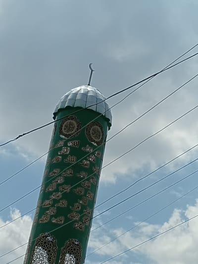 The image shows a close-up, low-angle shot of a minaret in the city of Osh, Kyrgyzstan. The minaret is cylindrical and painted green, adorned with intricate gold Arabic calligraphy and decorative patterns. It is topped with a silver, dome-like structure with a crescent moon finial. Several dark power lines crisscross the frame in front of the minaret and the sky. The sky is partly cloudy with patches of blue visible. The minaret appears to be part of a mosque, a prominent religious structure in Osh. The calligraphy on the minaret is likely verses from the Quran or the names of Allah.