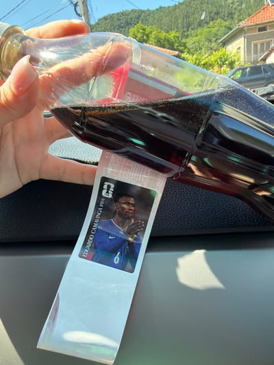 In the city of Beli Osam, Bulgaria, a hand is holding a clear glass bottle filled with a dark liquid. A partially visible sticker is attached to the bottle, featuring an image of a man in a blue soccer jersey with the number 6. The background shows a sunny day with green trees and houses, indicating an outdoor setting.