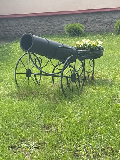 A black metal cannon replica, serving as a planter, sits on a green grassy area in front of a stone wall. The cannon has large spoked wheels. A basket of white flowers is attached to the side of the cannon. The background shows a stone wall with a building above it and some green bushes. The scene is located in the village of Kapyľski sielski savec, Belarus.
