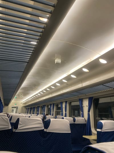 The interior of a passenger train car, consistent with railway services in China, is depicted, potentially operating within or connected to Huludao City. The scene shows rows of blue patterned seats with white headrest covers, configured in pairs along a central aisle and facing a single direction. The ceiling is white, arched, and illuminated by a central linear LED strip and multiple oval-shaped lights embedded in the side panels. A prominent blue louvered structure runs centrally along the ceiling, likely for ventilation, and similar blue louvered panels function as overhead luggage racks above the windows. Each window features blue curtains tied back. A small, white, square-shaped device is mounted on the reflective ceiling. Towards the rear of the car, a green illuminated emergency exit sign and a dark-colored dome surveillance camera are visible above a doorway. One individual is partially visible, obscured by a seat in the mid-ground on the right, clad in dark attire. The car appears largely empty. No discernible actions or interactions are occurring.