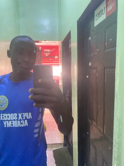 A selfie photograph shows a man wearing a blue soccer jersey with "Apex Soccer Academy" written on it. The photo seems to be taken indoors, with a door labeled "Ladies" visible in the background. The room appears to be simple, with green walls. While the location is reported as Tukur Tukur, Nigeria, there are no specific elements in the image that would verify this geographic information.