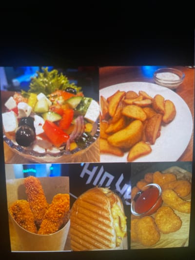 The image displays a collage of various food items. In the top left is a bowl of Greek salad with feta cheese, cucumbers, tomatoes, olives, and lettuce. To its right is a plate of golden potato wedges served with a small bowl of white sauce. The bottom left corner features a paper cone filled with three breaded chicken fingers. Next to it is a grilled sandwich. The bottom right shows a wooden board with chicken nuggets and a small dish of ketchup. The background of the bottom section features text that appears to be "HI N U" in large white letters. There is no indication of the location being in Paczkowo, Poland.