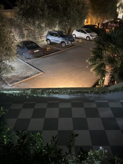 The image depicts an outdoor scene at night, likely in Trap, Montenegro. Several cars are parked on a paved surface. To the left, a white car is visible, followed by a dark gray car. Further down the row, another dark gray car with a license plate that appears to read "A598 CP 18" is parked. A yellow parking bollard is also visible.
The setting includes a paved area with a checkered pattern on a wall or structure to the right, composed of alternating dark and light squares. Lush greenery, including palm trees and other foliage, surrounds the parking area and part of the checkered structure. The lighting suggests it is nighttime, with artificial lights illuminating parts of the scene, casting warm glows on the pavement. The overall atmosphere is calm and still. There are no people visible in the image.