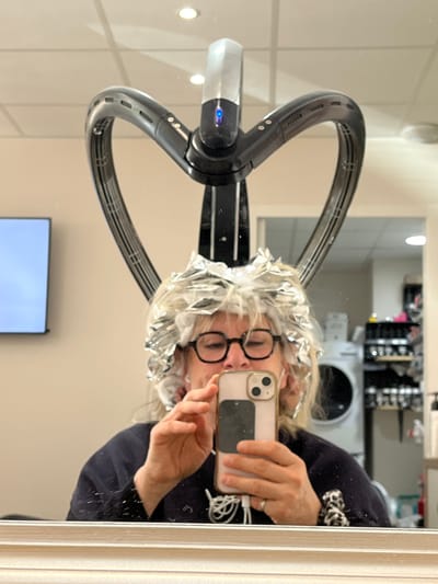 A woman with silver foil in her hair, possibly undergoing a hair treatment or coloring process, is reflected in a mirror. She is wearing glasses and holding a smartphone, seemingly taking a selfie. A large, black hair dryer or styling device is positioned over her head. The reflection reveals a salon setting with equipment and products visible in the background. A television screen is also reflected in the mirror. The location is assumed to be a hair salon in Montrond-les-Bains, France.