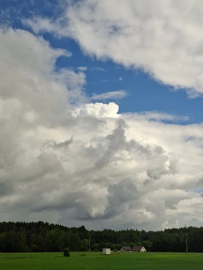 The image captures a rural landscape under a dramatic sky. In the foreground, a vibrant green field stretches out, dotted with a few small trees and a white cubic structure on the left. Further back, a line of dense, dark green forest forms the horizon, interrupted by two modest houses with tiled roofs and a single white structure. The sky above is a striking contrast of bright blue patches and voluminous white and grey clouds, with a particularly large, billowy cloud formation dominating the center. The scene suggests a day with dynamic weather, possibly transitioning between sun and rain, in a tranquil, undeveloped area. The location is identified as Balbieriškis, Lithuania.